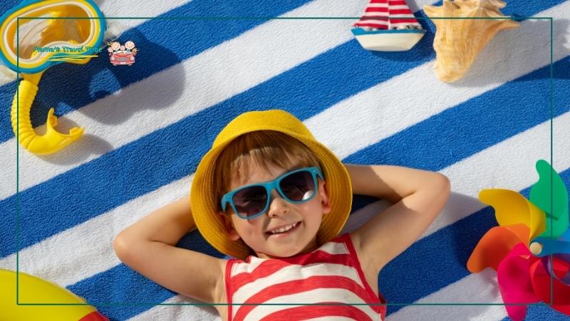 child lying down on a beach towel with sunglasses and a hat, surrounded by beach toys. toddler beach essentials