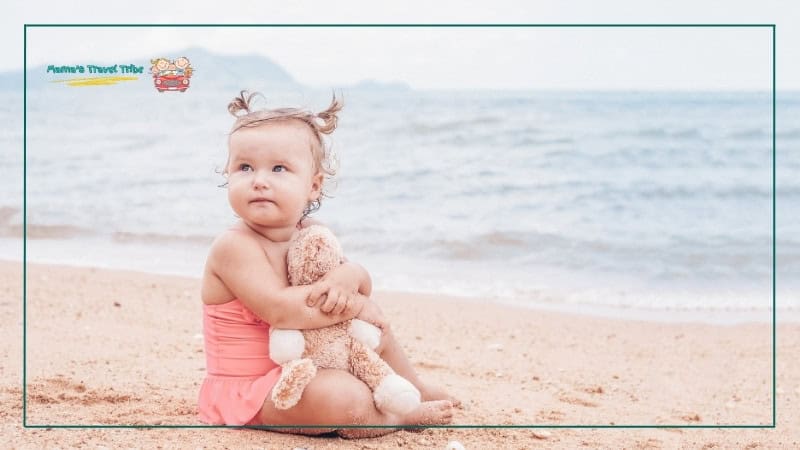 1 year old child sitting on the beach holding a teddy bear. simple beach days. best trips with toddlers. Trips and Activities by Age: Where to Go With Babies, Toddlers, and Preschoolers